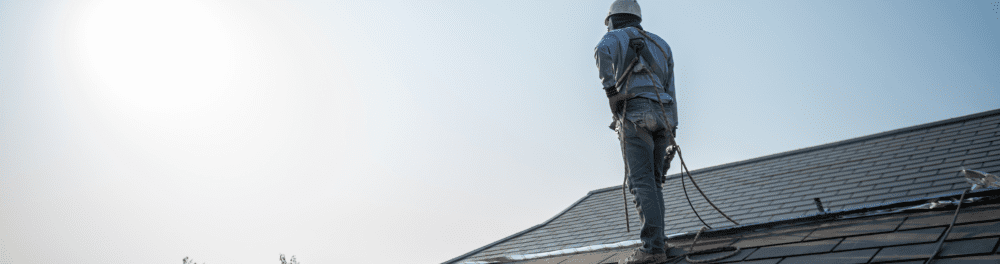 A roofer is shone standing on a roof in full safety gear. He is looking for signs of wear or damage.