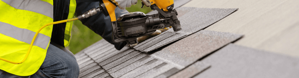 A roofer uses a nail gun to attach asphalt shingles to a sloped roof.